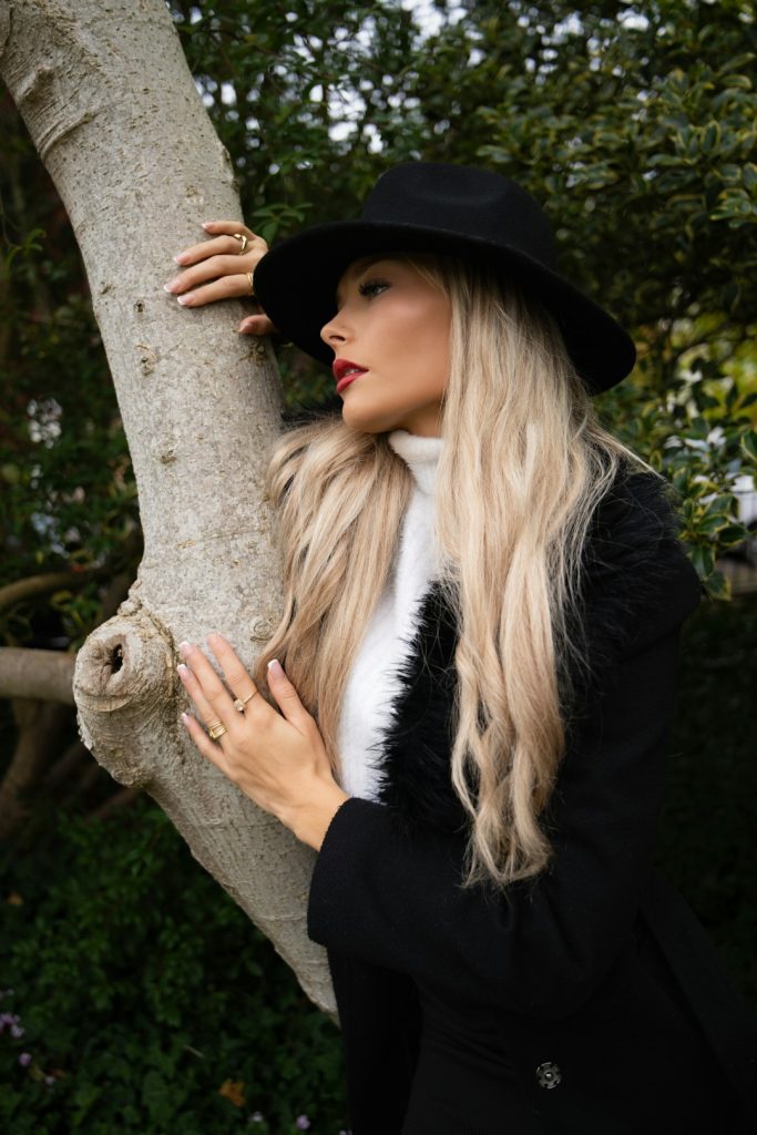 blonde-woman-in-black-hat-and-coat-leans-on-tree-zhiib5jkzvu