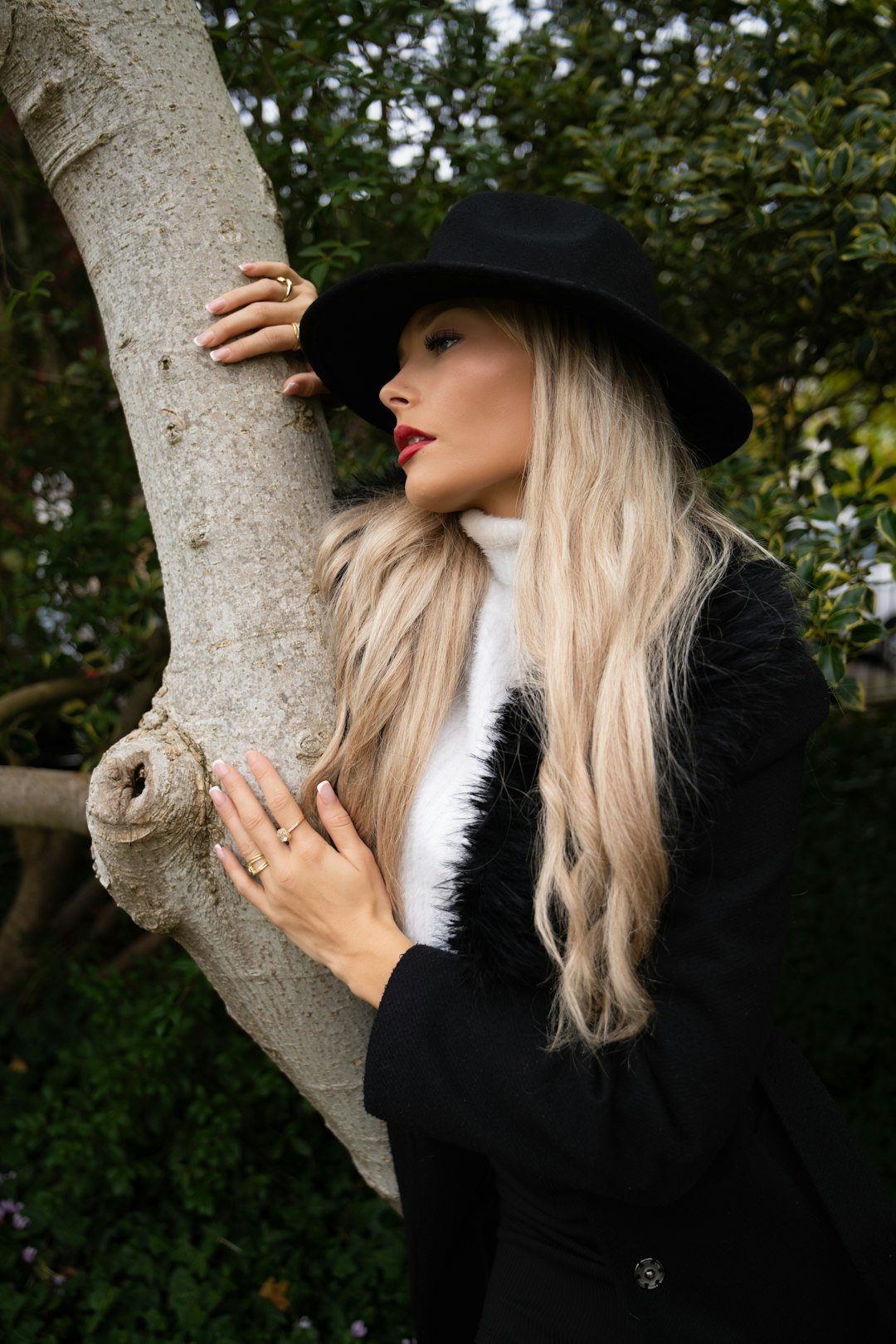 blonde-woman-in-black-hat-and-coat-leans-on-tree-zhiib5jkzvu